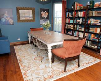 Dining table with two upholstered accent chairs and a set of six mid-century inspired transparent acrylic and chrome  chairs, wooden open frame bookcases, antique and vintage books, a large light color floral theme area rug, framed paintings and prints