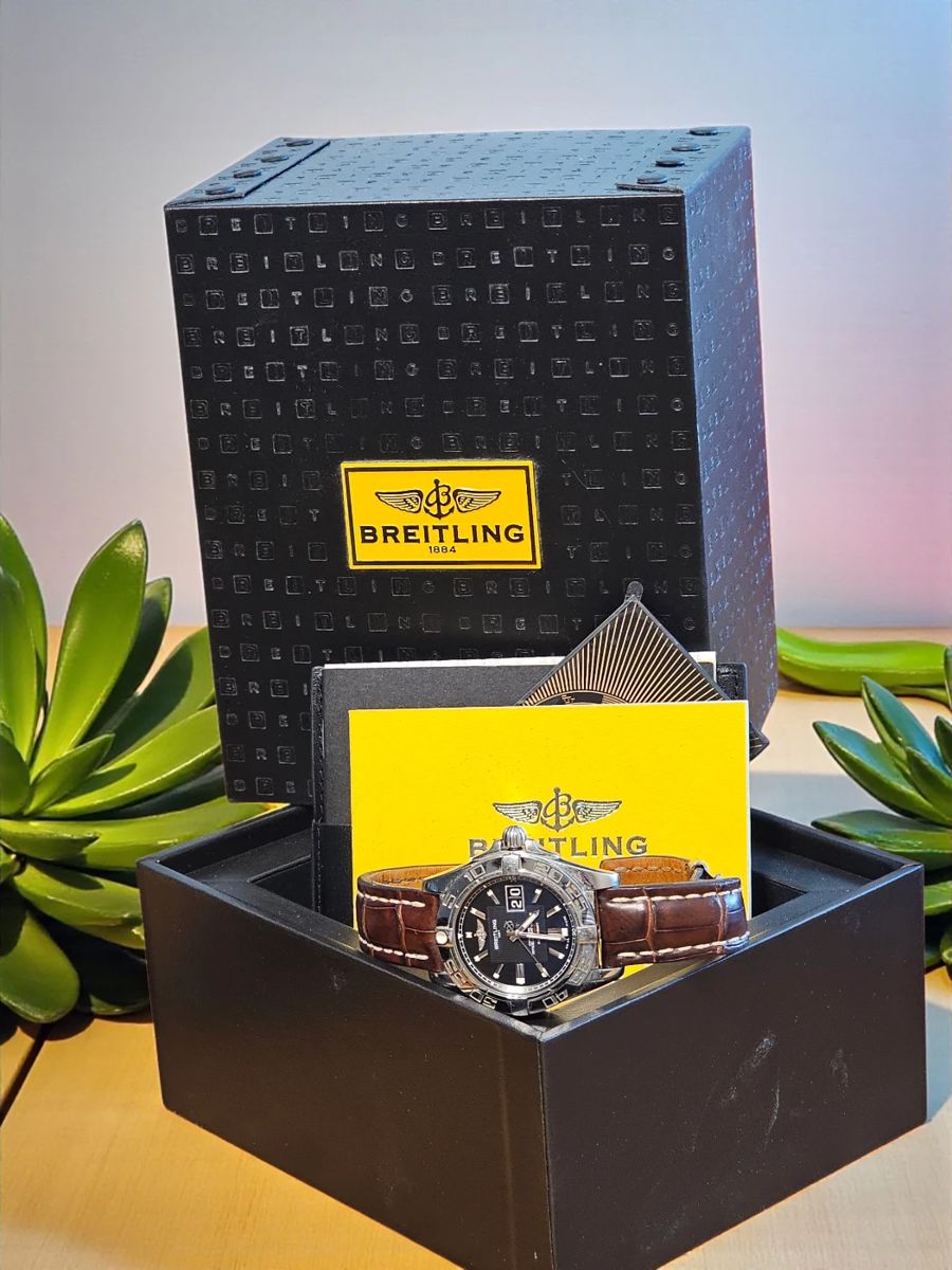Authentic Breitling men’s wristwatch featuring a black dial, date window, and rich brown alligator-style leather band. Includes original presentation box, warranty card, and booklet. Watch shows light surface wear consistent with gentle use.