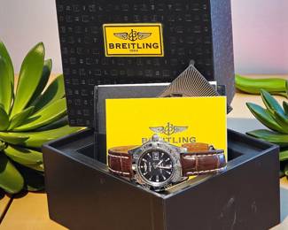 Authentic Breitling men’s wristwatch featuring a black dial, date window, and rich brown alligator-style leather band. Includes original presentation box, warranty card, and booklet. Watch shows light surface wear consistent with gentle use.