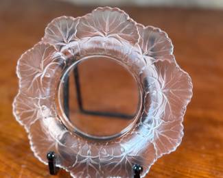 This lot features a Lalique "Honfleur" crystal dish, signed Lalique as shown in photos and measures, 5.75” H x 5.75” W. The design features a frosted geranium leaf pattern around the rim with a clear, glossy finish in the center. It is part of the "Honfleur" pattern, which is known for its geranium leaf design.
