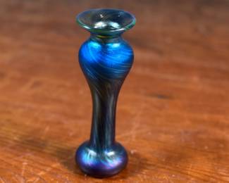 This lot features an iridescent vase by Orient & Flume. This vase measures, 5.5” H x 2” W. Signed underneath as shown in photos with original Orient & Flume sticker. Orient & Flume is an American art glass studio founded in 1972 in Chico, California. The name of the studio comes from the streets where its first location was, between Orient Street and Flume Street. The company is known for producing fine art glass pieces, including vases, paperweights, perfume bottles, and bowls. Their work is characterized by iridescent glass and clear crystal glass with intricate lampwork designs, often inspired by nature.
