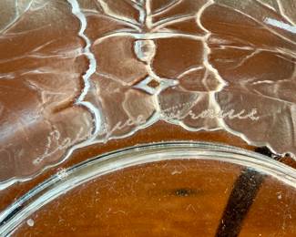 This lot features a Lalique "Honfleur" crystal dish, signed Lalique as shown in photos and measures, 5.75” H x 5.75” W. The design features a frosted geranium leaf pattern around the rim with a clear, glossy finish in the center. It is part of the "Honfleur" pattern, which is known for its geranium leaf design.
