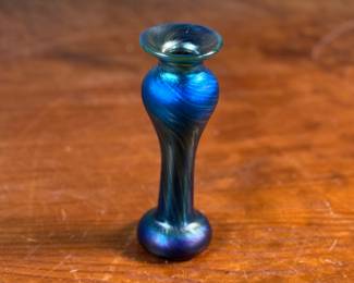 This lot features an iridescent vase by Orient & Flume. This vase measures, 5.5” H x 2” W. Signed underneath as shown in photos with original Orient & Flume sticker. Orient & Flume is an American art glass studio founded in 1972 in Chico, California. The name of the studio comes from the streets where its first location was, between Orient Street and Flume Street. The company is known for producing fine art glass pieces, including vases, paperweights, perfume bottles, and bowls. Their work is characterized by iridescent glass and clear crystal glass with intricate lampwork designs, often inspired by nature.
