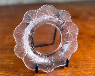 This lot features a Lalique "Honfleur" crystal dish, signed Lalique as shown in photos and measures, 5.75” H x 5.75” W. The design features a frosted geranium leaf pattern around the rim with a clear, glossy finish in the center. It is part of the "Honfleur" pattern, which is known for its geranium leaf design.
