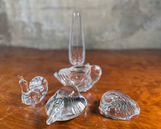 This lot features a collection of four (4) animal themed Steuben Glass sculptures along with one tall skinny Steuben Glass vase, with with Steuben sleeves to accompany them. Steuben Glass is an American art glass company founded in Corning, New York, in 1903 by Frederick Carder. Originally known for colored glass, it shifted to brilliant clear crystal in the 1930s after being acquired by Corning Glass Works (now Corning Inc.) in 1918. The brand was sold in 2008, production ceased in 2011, and was reacquired by Corning Inc., which now oversees its production and sales through the Corning Museum of Glass.
