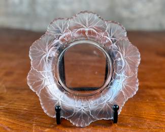This lot features a Lalique "Honfleur" crystal dish, signed Lalique as shown in photos and measures, 5.75” H x 5.75” W. The design features a frosted geranium leaf pattern around the rim with a clear, glossy finish in the center. It is part of the "Honfleur" pattern, which is known for its geranium leaf design.
