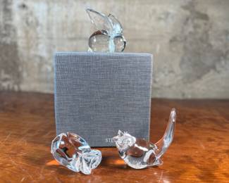 This lot features a collection of three (3) animal themed Steuben Glass sculptures, with five cases to accompany them. Steuben Glass is an American art glass company founded in Corning, New York, in 1903 by Frederick Carder. Originally known for colored glass, it shifted to brilliant clear crystal in the 1930s after being acquired by Corning Glass Works (now Corning Inc.) in 1918. The brand was sold in 2008, production ceased in 2011, and was reacquired by Corning Inc., which now oversees its production and sales through the Corning Museum of Glass.
