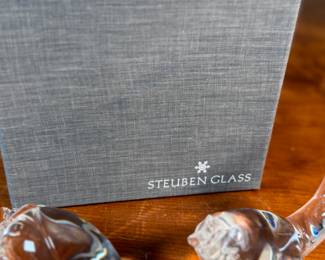 This lot features a collection of three (3) animal themed Steuben Glass sculptures, with five cases to accompany them. Steuben Glass is an American art glass company founded in Corning, New York, in 1903 by Frederick Carder. Originally known for colored glass, it shifted to brilliant clear crystal in the 1930s after being acquired by Corning Glass Works (now Corning Inc.) in 1918. The brand was sold in 2008, production ceased in 2011, and was reacquired by Corning Inc., which now oversees its production and sales through the Corning Museum of Glass.
