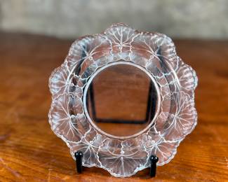 This lot features a Lalique "Honfleur" crystal dish, signed Lalique as shown in photos and measures, 5.75” H x 5.75” W. The design features a frosted geranium leaf pattern around the rim with a clear, glossy finish in the center. It is part of the "Honfleur" pattern, which is known for its geranium leaf design.
