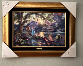 Framed print titled 'The Princess and the Frog' by Thomas Kinkade, featuring a colorful scene with characters near water lilies and a riverboat. The frame is gold with ornate detailing and includes a small engraved plaque with the title and artist's name. The canvas size is 18 by 27 inches, with print number 52310 of 695. The print is signed by Thomas Kinkade. The certificate of limitation and authenticity is also visible, printed on white paper with black text.