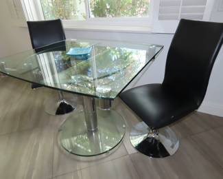 Square glass table extends to oval table, two black leather swivel dining chairs.