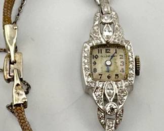 Antique diamond 17 jewel ladies watch (currently not working) is marked Ollendorff, a NY company that imported Swiss watches. Diamonds are around watch face and form half of the band. Safety chain is attached to watch. Watch is marked 900 Platinum .100 Iridium. Watch is engraved on back; 5.5"L, 13.50g (includes partial cloth band). Band is marked 14kt as shown in photos