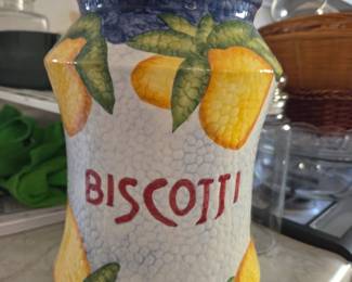 A tall, lidded ceramic jar labeled "Biscotti" in bold red letters. Features a textured white background with hand-painted yellow fruit and green leaves, accented by deep blue trim along the top and bottom. A colorful, decorative piece ideal for storing cookies or adding charm to a kitchen counter.