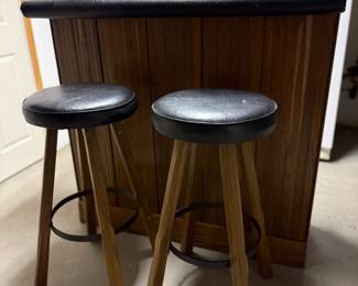 Compact vintage home bar and matching stools. The bar features warm wood paneling, a black padded vinyl top, and two coordinating barstools with sturdy wood legs, black vinyl seats, and metal footrests.