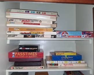 games visible include:
Top shelf: The Game of Life, The Mad Magazine Game, Monopoly, Parcheesi, Bonkers!, and Ouija.
Bottom shelf: The Big Bang Theory Fact or Fiction Trivia Game, Passtimes: The Game of History, Tripoley, Topy Color, Geografacts, and Aggravation.