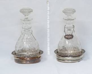 A pair of antique cut-glass decanters with stoppers, possibly Georgian or Regency period, sitting in silver-plated coasters. Both decanters have wheel cut decoration of fruiting grape vines, and one bears an ornate sterling silver label on linked chain, reading "Bourbon" and on the reverse, stamped sterling and W for makers mark. 
