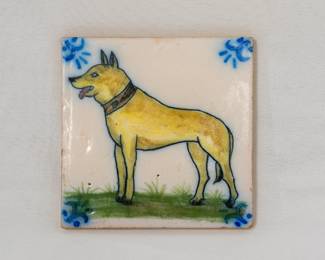 A hand-painted ceramic tile featuring a dog, with blue floral motifs in the corners and green grass at the bottom; crafted in Portugal