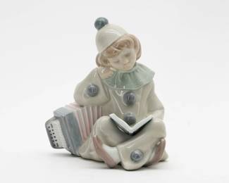 From Lladro’s Pierrot Children series, this porcelain figurine is of a seated girl in Pierrot costume, leaning on an accordion while she reads a book, and has a clear shiny glaze. Measuring 5 inches H. Small chip to left side of book cover. The Pierrot Children series from Lladro is from 1971–1982. Pierrot is a clown character from the Italian commedia dell'arte.
