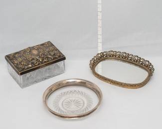 A vintage glass trinket box with an ornate, embossed floral and scrollwork silver plated hallmarked lid, a sterling silver and glass coaster or trinket dish, and a vintage gold-toned mirrored vanity tray with an ornate pierced metal rim