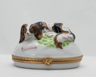 A hand-painted Limoges porcelain trinket box with a Cavalier King Charles Spaniel reclining on the lid near a pink dog bone, the box and lid with brass band and flower-shaped clasp. Marked to the base with Limoges France and Tiffany & Co, showing this was made in the Limoges region of France and sold by Tiffany & Co.