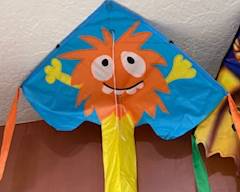 Bright and colorful kids’ kite featuring a cheerful orange monster character with long streaming tails—fun outdoor toy for parks, beaches, and backyard play.