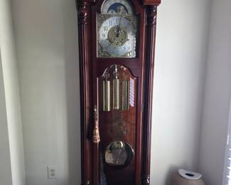 This Sligh grandfather clock is a floor-standing timepiece crafted from rich, polished hardwood, likely cherry or mahogany, with a warm reddish-brown finish. The clock features a detailed dial with Arabic numerals, ornate filigree accents, and a moon phase complication on the upper left. The pendulum and chimes are visible through a glass panel on the right side of the case. The overall design suggests a traditional or classic style, suitable for a formal living space or study. The clock appears to be in good condition, with a polished surface and clear glass. Client tells the story that it was the last Sligh to be made.