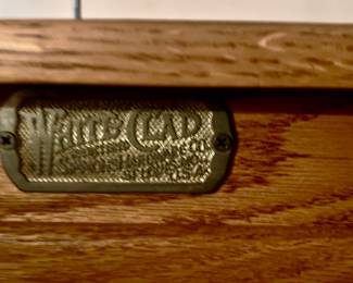 The image shows a metal nameplate from a "White Clad" cabinet or ice box, a trademark of the Simmons Hardware Company. The company operated from 1874 to 1939 and produced various oak furniture pieces in the Mission/Arts and Crafts style.