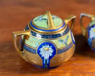 This lot features an antique sugar bowl with lid and a cream pitcher set, hand-painted in an Art Nouveau style by an artist named Osborne for Pickard China. The set features a design with stylized flowers on a cobalt ground and multiple borders of blue and white luster colors.