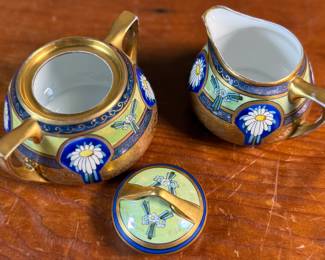 This lot features an antique sugar bowl with lid and a cream pitcher set, hand-painted in an Art Nouveau style by an artist named Osborne for Pickard China. The set features a design with stylized flowers on a cobalt ground and multiple borders of blue and white luster colors.