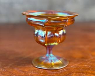 This lot features an iridescent gold Tiffany Favrile sherbet glass with maker marks present underneath as shown in photos. One small repair on the foot ring and measures, 3.5” H x 4” W x 4” D.