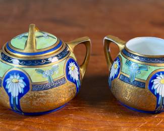 This lot features an antique sugar bowl with lid and a cream pitcher set, hand-painted in an Art Nouveau style by an artist named Osborne for Pickard China. The set features a design with stylized flowers on a cobalt ground and multiple borders of blue and white luster colors.