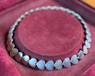 This lot features an iconic sterling silver heart necklace titled, "Chain of Hearts". A Hans Hansen design for Georg Jensen with a total approx. weight of 61.77 grams and measures, 15.5”. Comes in the original box and in excellent condition. The design features a series of meticulously crafted, solid silver heart shapes linked together to create an articulated, choker-style necklace. Maker marks present as shown in photos.