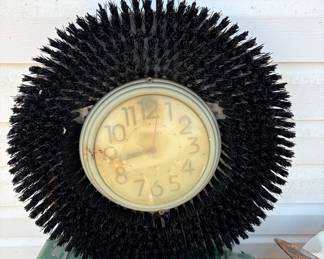  Old analog clock face mounted within a large, used floor scrubber brush. 
