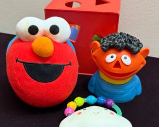 Children’s toys, including a plush Elmo doll, an Ernie pop-up toy or rattle, a red shape-sorter box, and a small teether/rattle toy.