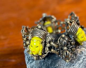 This lot features an unmarked "Asian Princess" bracelet in the style of Selro Selini. This bracelet features yellow molded plastic faces set in ornate, dark metal links. Measures 5-6” depending on use of safety chain. 
