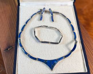 This lot features a vintage silver lapis modernist set including a hinged bib necklace, a bracelet, and earrings. Marked .970 silver with a total approx. weight of 51.89 grams. The necklace measures 16”, bracelet 8”, and earrings 1.5” H.
