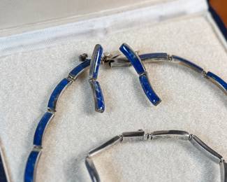 This lot features a vintage silver lapis modernist set including a hinged bib necklace, a bracelet, and earrings. Marked .970 silver with a total approx. weight of 51.89 grams. The necklace measures 16”, bracelet 8”, and earrings 1.5” H.

