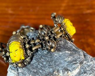 This lot features an unmarked "Asian Princess" bracelet in the style of Selro Selini. This bracelet features yellow molded plastic faces set in ornate, dark metal links. Measures 5-6” depending on use of safety chain. 
