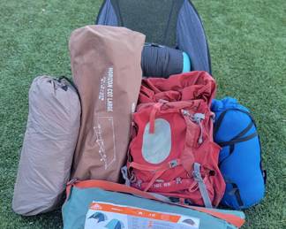 Camping gear including two Kelty tents, REI camp cot, Osprey women's backpack, sleeping bags with compression bags, camp chairs, additional camping gear and supplies.