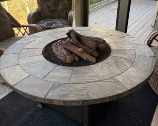 Large round propane fire pit table with stone-look tile top and faux log set, on sturdy metal base.