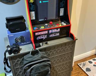 Canon PowerShot digital camera with bag. Additionally, there is a travel backpack specifically designed for cameras. Furthermore, there is a GoPro series 4 with two additional batteries. Lastly, there is a tabletop video game Pac-Man, along with three other games, built into the device.