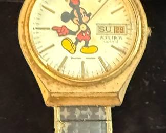 vintage Bulova Accutron Mickey Mouse watch.
It features a day and date display, currently showing "SU 28".
The watch likely dates back to the 1970s.
It has the classic Mickey Mouse character on the dial, with his arms
