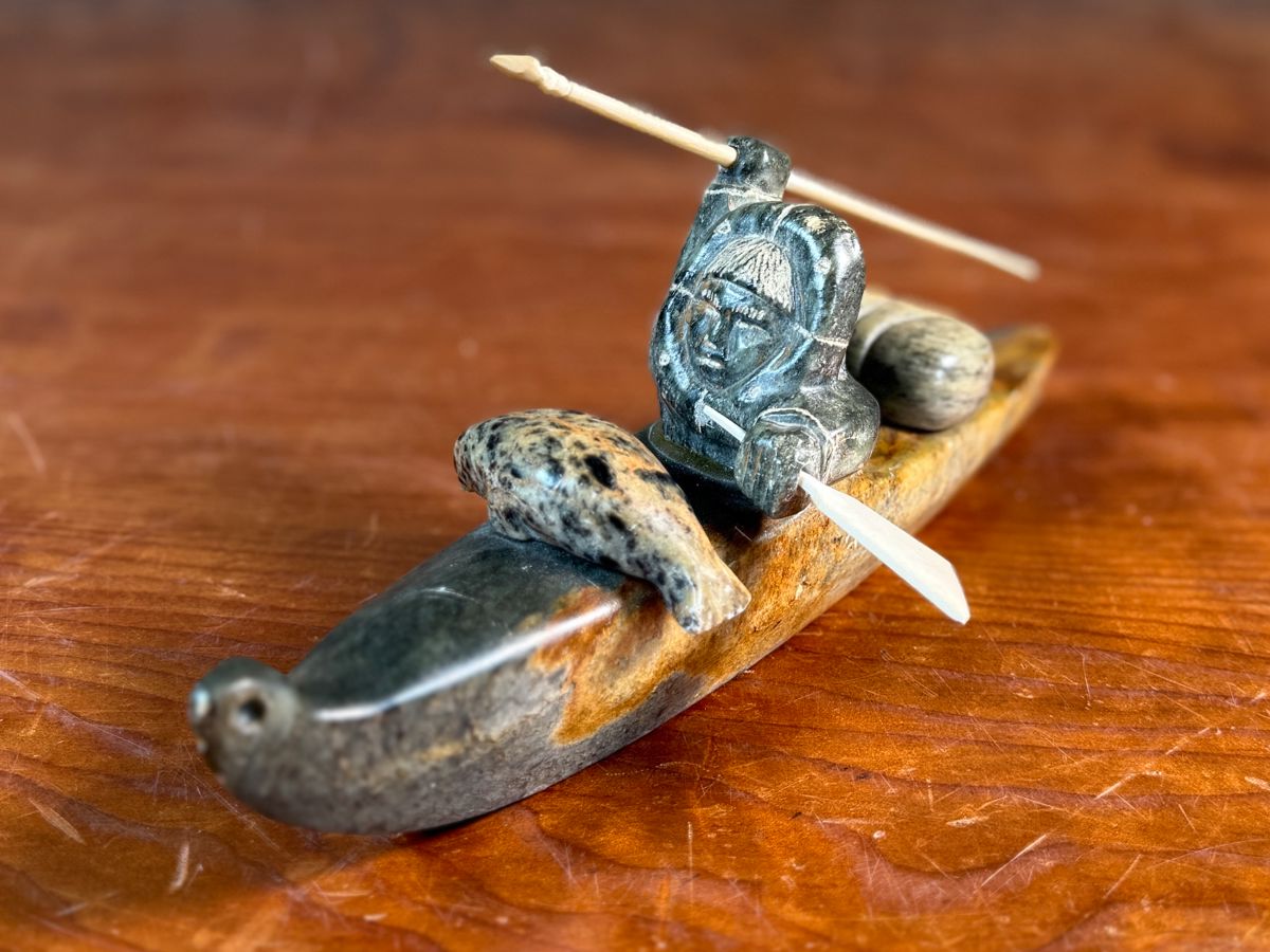 "Hunter and Kayak", an Inuit hand-carved sculpture, likely made of soapstone by listed artist, Arnold Kayutak. Signed underneath as shown in photos and measures, 4” H x 9.5” W x 4” D.

