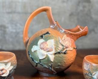 This lot features a collection of seven (7) Roseville Pottery items in the "Magnolia" pattern, which was introduced in 1943. The set includes a large cider pitcher and several matching mugs, featuring pink and white blossoms on a textured russet and green ground. All items are in excellent condition with one piece showing a very small chip as shown in photos. The pitcher measures, 8.5” H.
