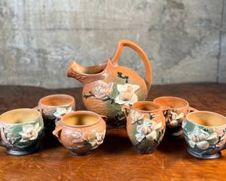 This lot features a collection of seven (7) Roseville Pottery items in the "Magnolia" pattern, which was introduced in 1943. The set includes a large cider pitcher and several matching mugs, featuring pink and white blossoms on a textured russet and green ground. All items are in excellent condition with one piece showing a very small chip as shown in photos. The pitcher measures, 8.5” H.
