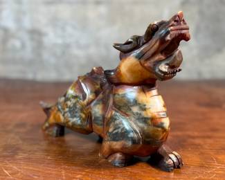 This lot features a beautiful ochre, copper, and viridescent example of a Chinese Pixiu. The creature is depicted with mouth agape, and standing in a fighting stance, his tail twitches behind him frozen in time. Pixiu is a Chinese mythical hybrid creature. Pixiu are considered powerful protectors of the souls of the dead, and resemble strong, winged lions. This sculpture of a Pixiu measures, 7” H x 3” W x 9” D.
