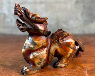 This lot features a beautiful ochre, copper, and viridescent example of a Chinese Pixiu. The creature is depicted with mouth agape, and standing in a fighting stance, his tail twitches behind him frozen in time. Pixiu is a Chinese mythical hybrid creature. Pixiu are considered powerful protectors of the souls of the dead, and resemble strong, winged lions. This sculpture of a Pixiu measures, 7” H x 3” W x 9” D.
