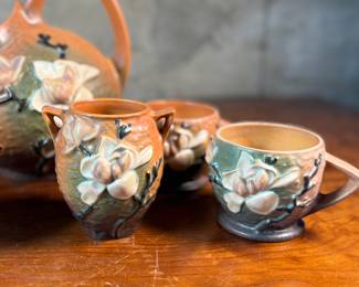 This lot features a collection of seven (7) Roseville Pottery items in the "Magnolia" pattern, which was introduced in 1943. The set includes a large cider pitcher and several matching mugs, featuring pink and white blossoms on a textured russet and green ground. All items are in excellent condition with one piece showing a very small chip as shown in photos. The pitcher measures, 8.5” H.
