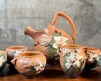This lot features a collection of seven (7) Roseville Pottery items in the "Magnolia" pattern, which was introduced in 1943. The set includes a large cider pitcher and several matching mugs, featuring pink and white blossoms on a textured russet and green ground. All items are in excellent condition with one piece showing a very small chip as shown in photos. The pitcher measures, 8.5” H.

