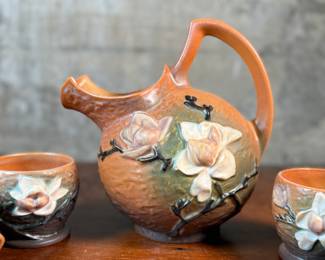 This lot features a collection of seven (7) Roseville Pottery items in the "Magnolia" pattern, which was introduced in 1943. The set includes a large cider pitcher and several matching mugs, featuring pink and white blossoms on a textured russet and green ground. All items are in excellent condition with one piece showing a very small chip as shown in photos. The pitcher measures, 8.5” H.
